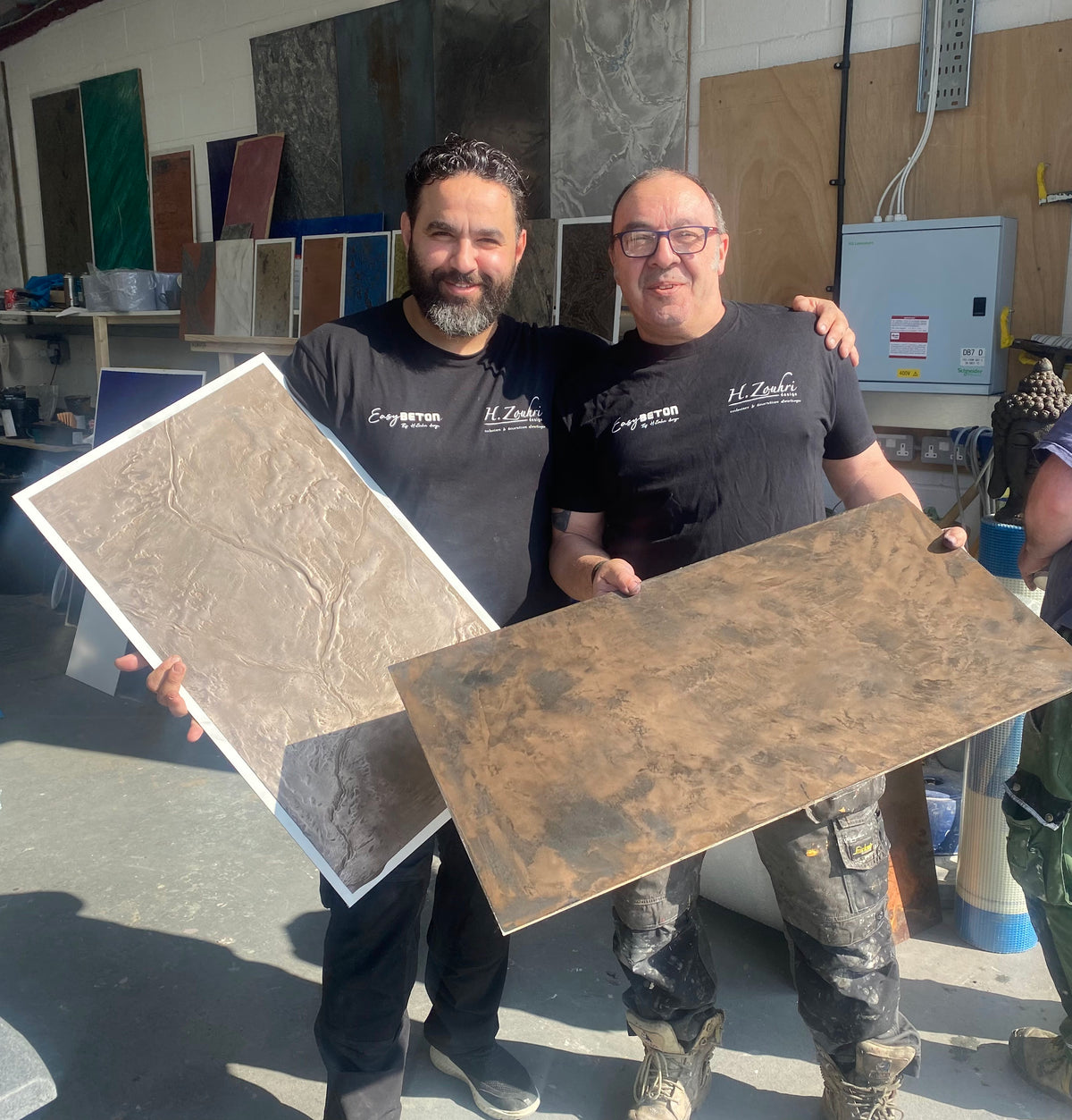 Two Microcement Company specialists holding finished decorative sample panels in the workshop